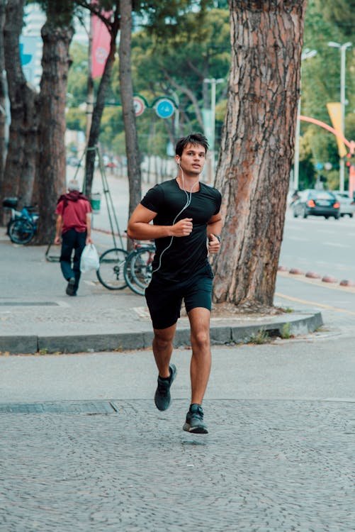 Free Fit man jogging with earphones on a tree-lined city street, promoting healthy lifestyle. Stock Photo