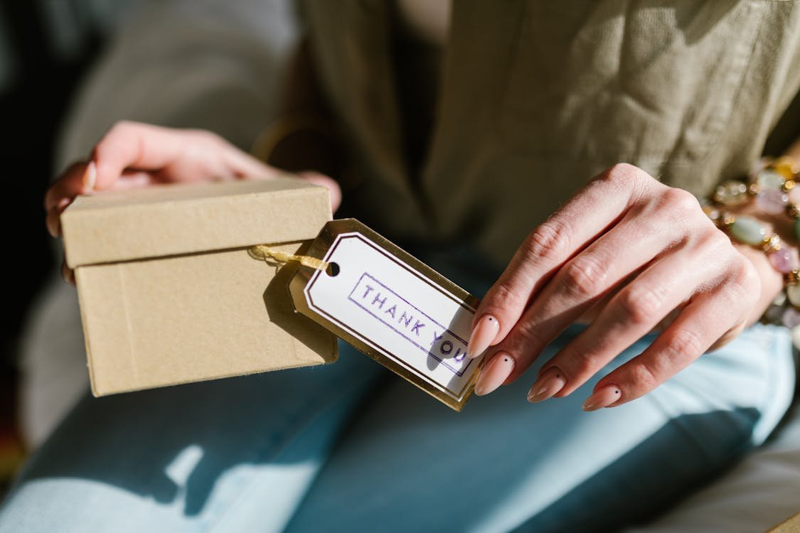 Free Close-up of hands holding a gift box with a Thank You tag attached, in natural lighting. Stock Photo