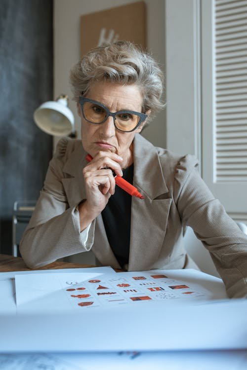 Free Senior woman wearing blazer, pensively reviewing documents at her workspace, holding a pen. Stock Photo