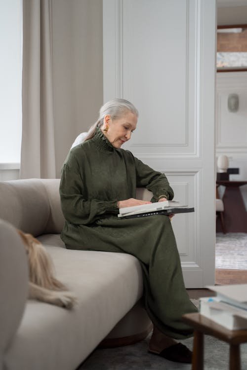 Free Elderly woman in a green dress reading on a sofa at home, creating a calm and cozy atmosphere. Stock Photo