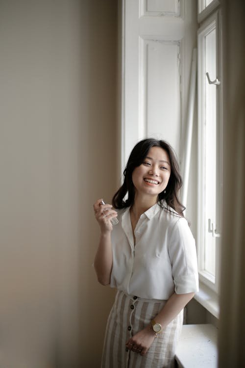 Free Happy Asian woman in casual clothes smiling and spraying aromatic perfume on neck while resting near window at home Stock Photo