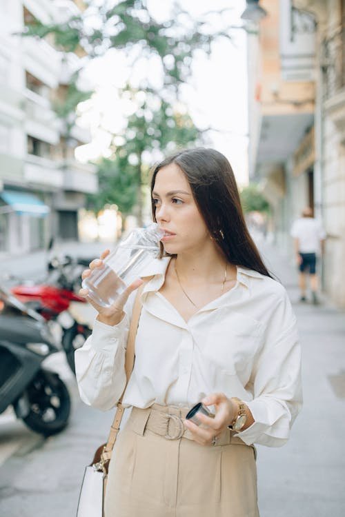 Free A stylish woman in white shirt sipping water on a sunny city street Stock Photo