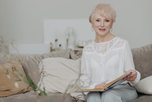 Free Senior woman with white hair sitting on a couch and reading a book with a warm smile. Stock Photo