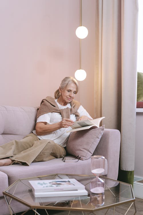 Free Elderly woman enjoying leisure time reading a book on a sofa in a cozy living room. Stock Photo